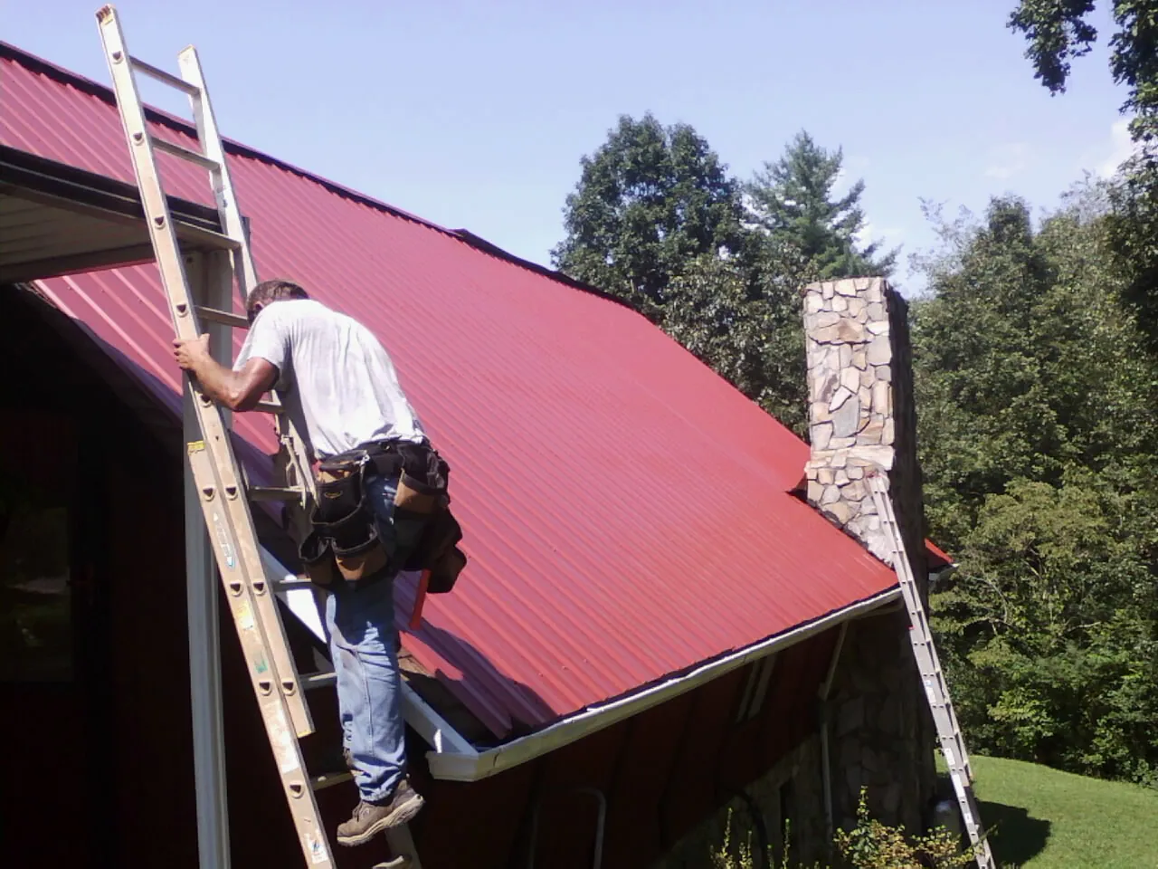 Roofer on a ladder beside a red metal roof and stone chimney — Soffit & Fascia Repair by Roofing Brothers in King Of Prussia